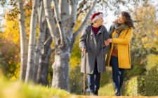 Granddaughter walking with senior woman in park wearing winter clothing. Old grandmother with walking cane walking with lovely caregiver girl in sunny day. Happy woman and smiling grandma walking in autumn park.